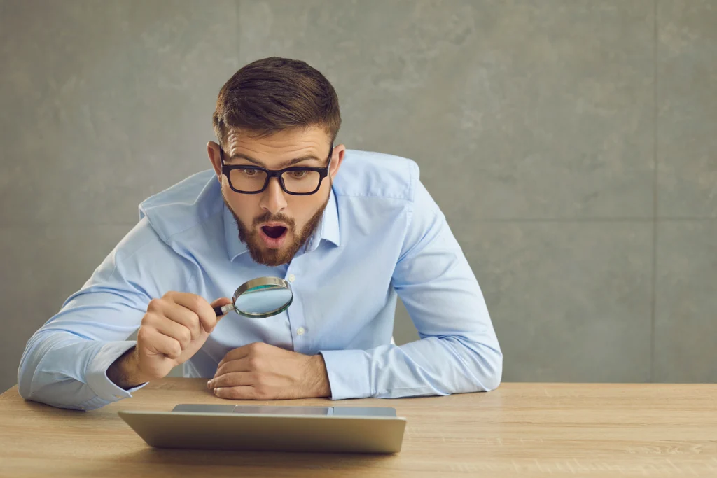 Jeune homme avec des lunettes examinant attentivement sa déclaration d’impôt sur un ordinateur à l’aide d’une loupe, illustrant la rectification d’impôt à Genève.