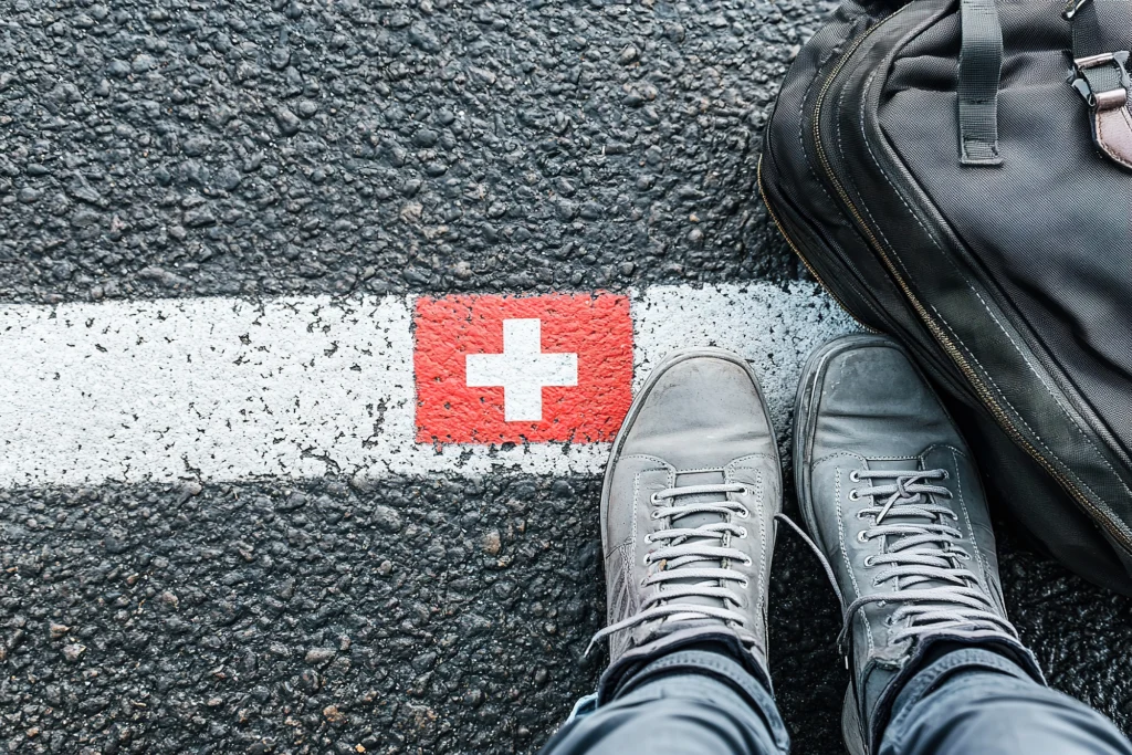 Travailler en Suisse et vivre en France : chaussures sur la ligne marquée d’un drapeau suisse à la frontière, illustration des impôts des frontaliers